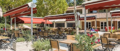 An outdoor cafe with wooden tables and chairs, covered by red awnings, and surrounded by planters.