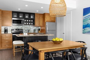 Cozinha moderna com armários de madeira, eletrodomésticos de aço inoxidável, sala de jantar com mesa e cadeiras de madeira, e luminária de vime suspensa.