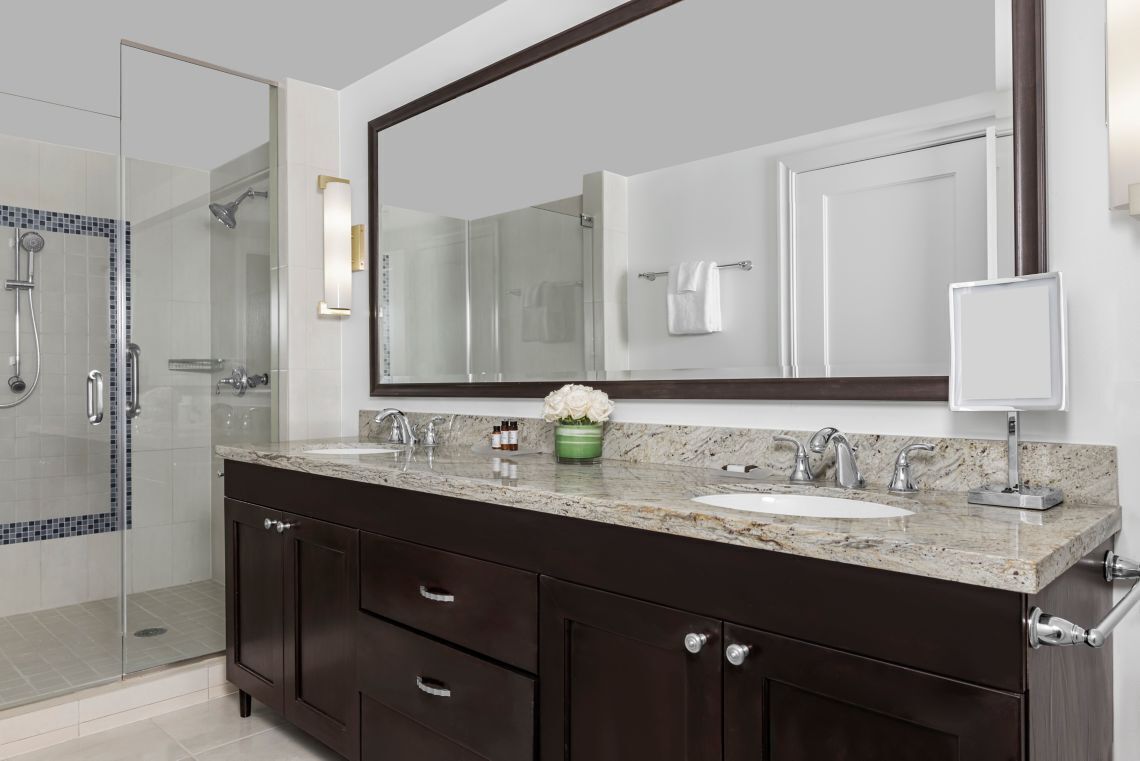 Un bagno moderno con cabina doccia in vetro, mobili scuri in legno, un piano di lavoro in marmo e uno specchio grande.