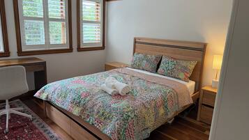 A bedroom with a wooden bed, a desk, a chair, and two windows with blinds.