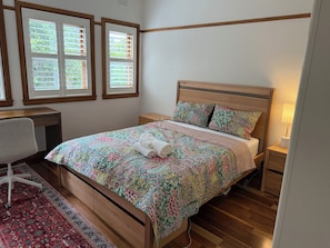 Ein Schlafzimmer mit einem hölzernen Bett, einem Schreibtisch, einem Stuhl und zwei Fenstern mit Jalousien.
