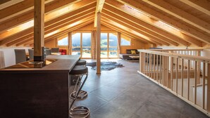 Une pièce mansardée spacieuse avec un plafond haut, une longue structure en poutres apparentes, une grande fenêtre offrant une vue sur les montagnes, un coin cuisine avec des tabourets de bar et un espace salon avec un canapé et une table basse.