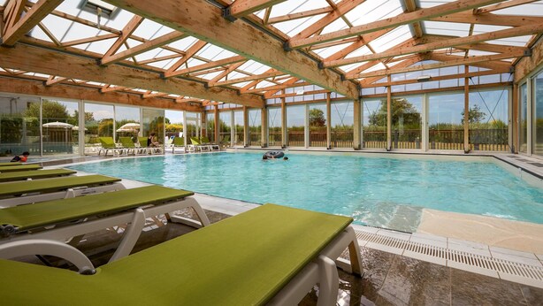 Piscina com vigas de madeira e paredes de vidro, rodeada por espreguiçadeiras.