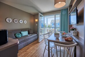 A modern living room with a sofa, dining table, chairs, a flat-screen TV, and artwork on the wall. - Courseulles Coastal Apartment for 4 (Courseulles-sur-Mer)