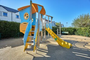 A playground with a slide and climbing structures, surrounded by a fence and a building in the background. - Courseulles Coastal Apartment for 4 (Courseulles-sur-Mer)