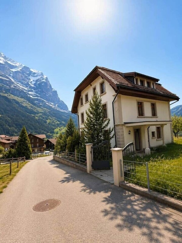 A two-story house with a sloped roof, a front porch, and a driveway leading to a gated entrance.