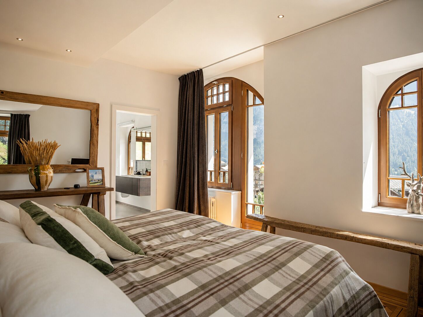 A bedroom with a large bed, a mirror, a window with curtains, and a view of the outdoors.