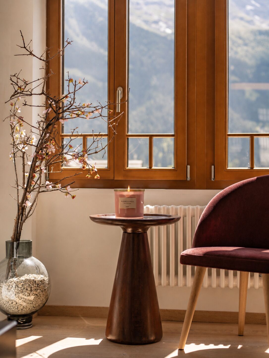 A room with a window, a vase with branches, a candle on a table, and a chair.