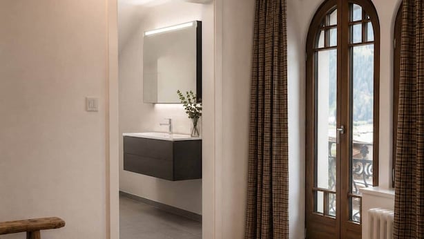 Una stanza con pavimento in legno, bagno moderno con specchio e lavandino, e vista attraverso una finestra con tende.