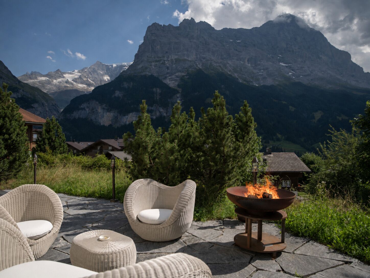 A patio with wicker furniture and a fire pit, overlooking a mountainous landscape.