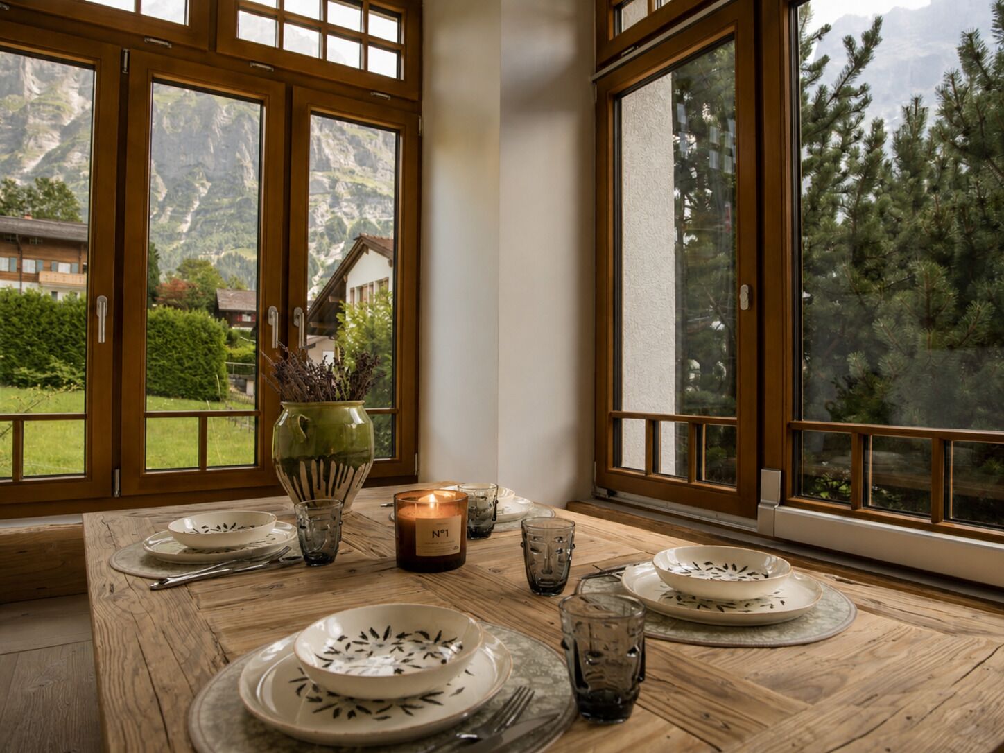 A dining table set with plates, glasses, and a candle, with a view of mountains through large windows.