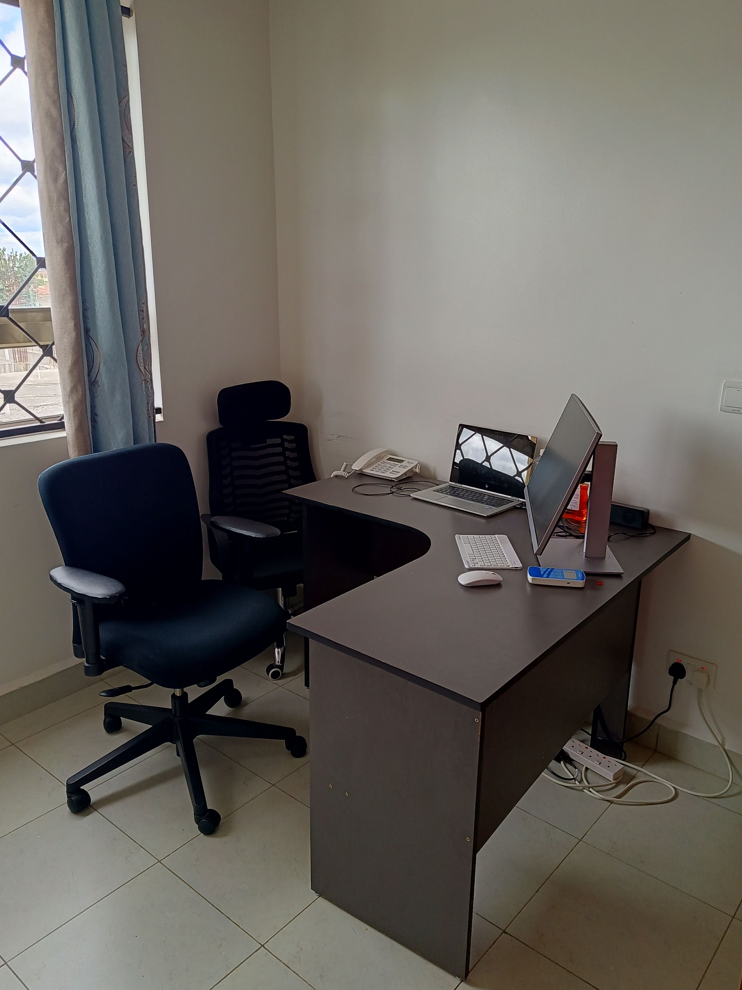 A desk with a laptop, a phone, and a computer monitor. There is a chair and a window with curtains.
