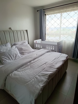 A bedroom with a large bed, a crib, a window with curtains, and a view of the outdoors. - Fantastic 2-bedroom apartment in tranquil Pridelands with AC (Pridelands)