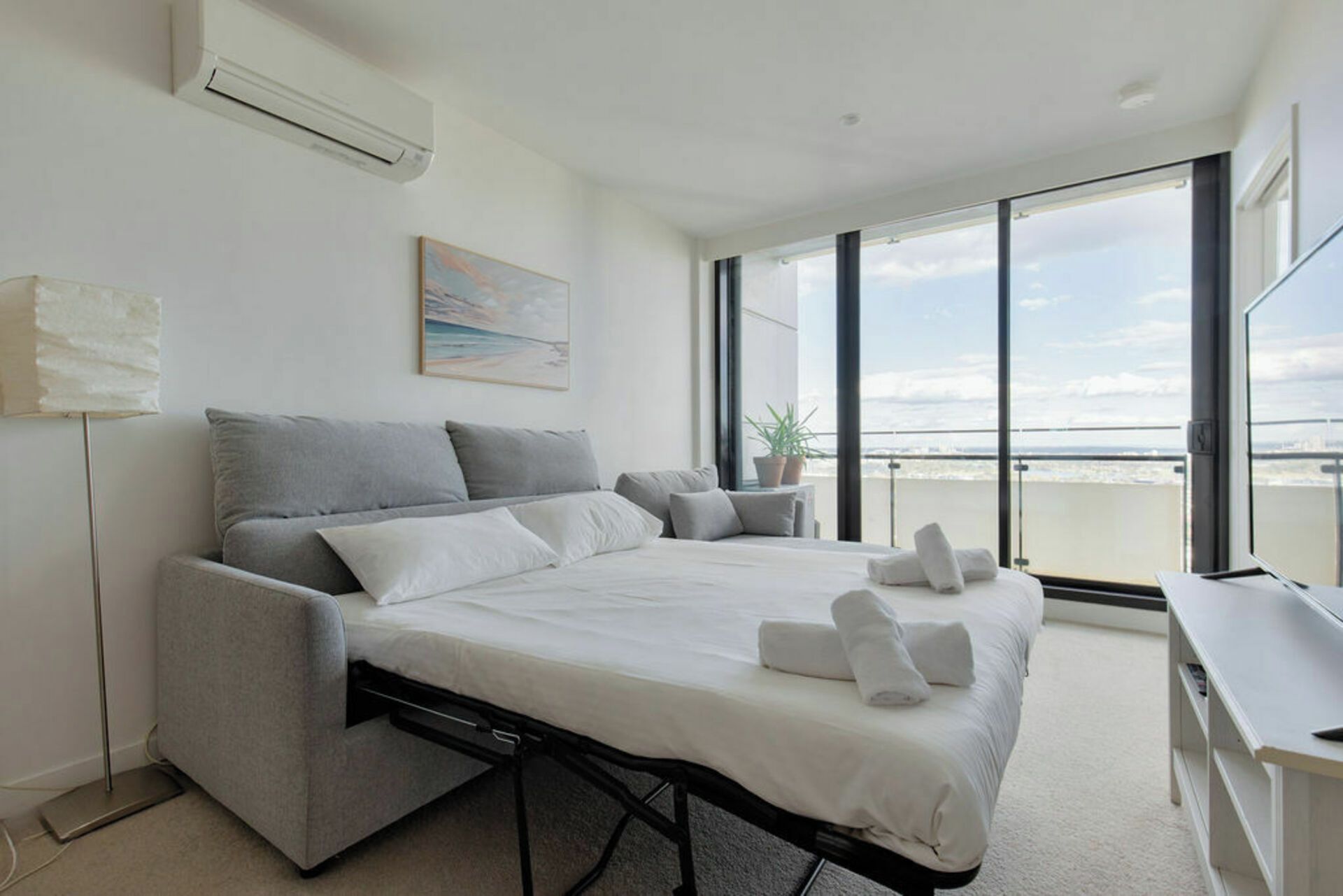 A modern bedroom with a bed, a desk, a potted plant, and a large window with a city view.