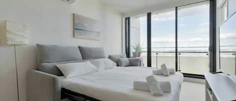A modern bedroom with a bed, a desk, a potted plant, and a large window with a city view.