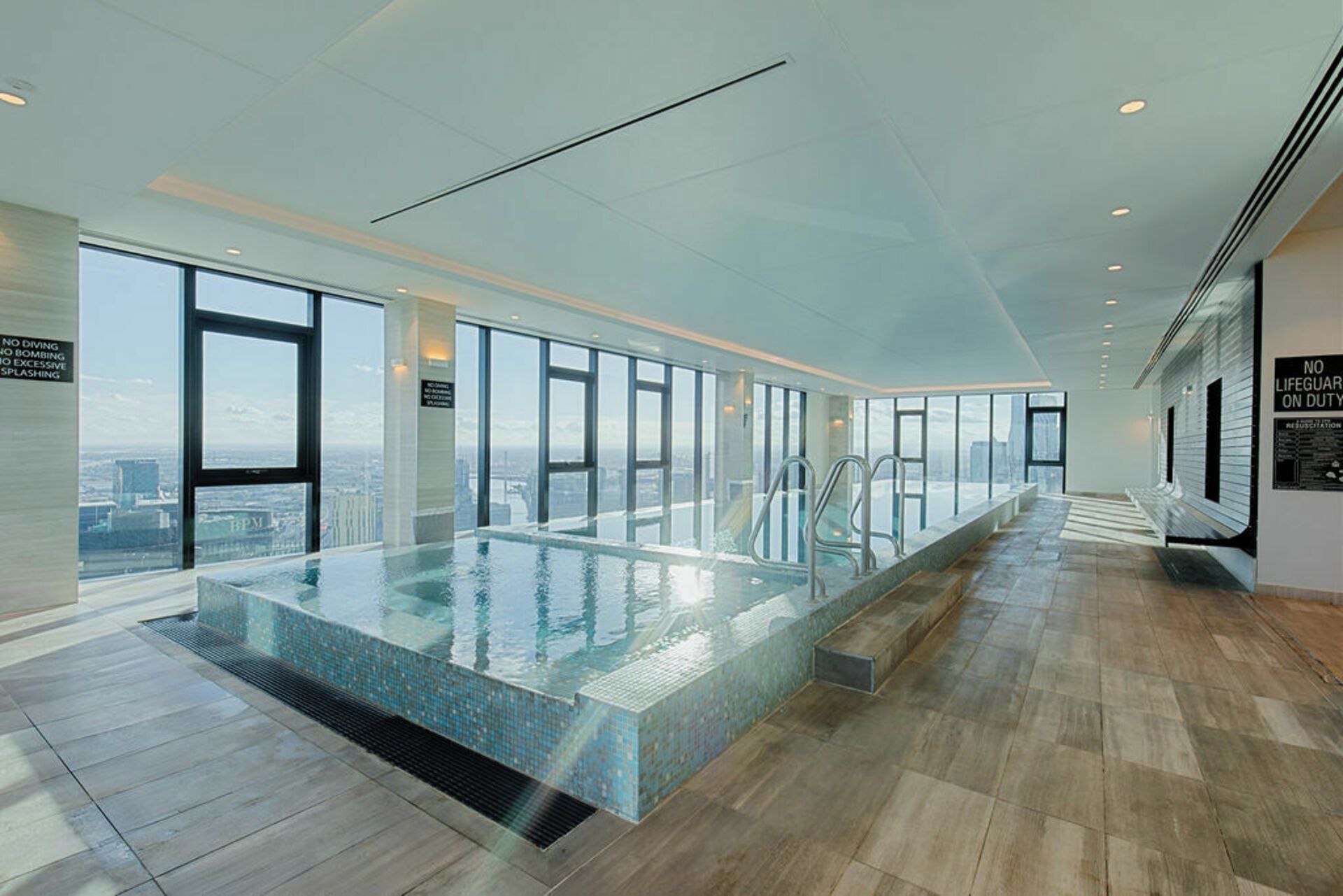 A modern indoor swimming pool with a city view, large windows, and a tiled floor.