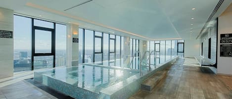 A modern indoor swimming pool with a city view, large windows, and a tiled floor.