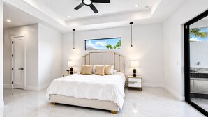 Un dormitorio moderno con una cama grande, dos mesitas de noche con lámparas, un ventilador de techo y un ventanal con vistas a árboles.