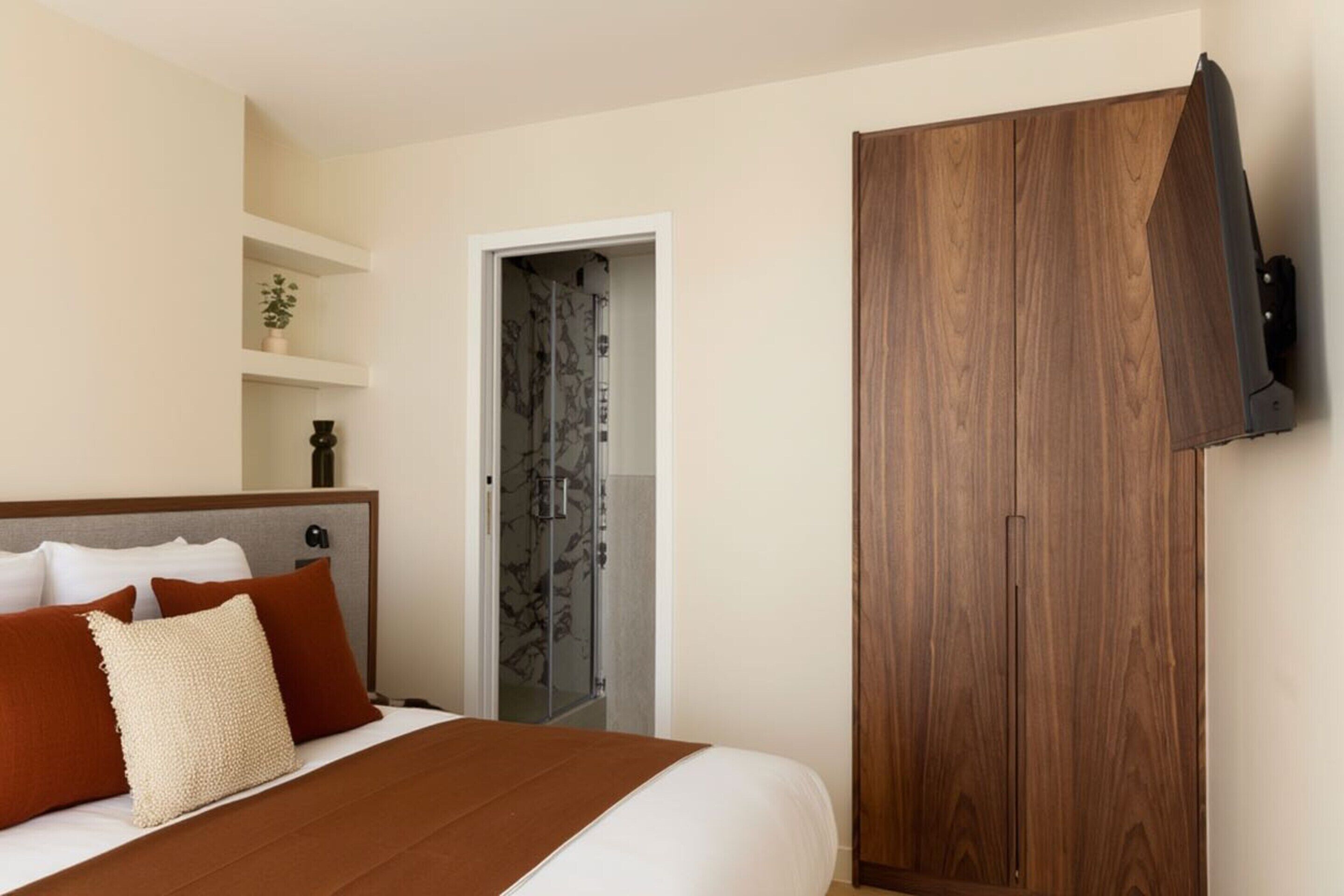 A bedroom with a bed, a wardrobe, a shelf with a plant, and a door leading to a bathroom.
