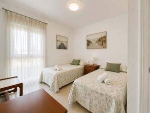 Ein Zimmer mit zwei Betten, einem kleinen Tisch, einem Nachttisch und einem Fenster mit durchsichtigen Vorhängen.