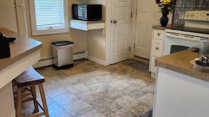 A kitchen with white cabinets, a microwave, a stove, and a sink. There is a window with blinds, a trash can, and a wooden stool.