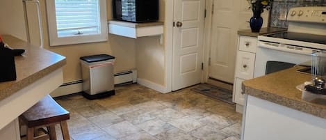 A kitchen with white cabinets, a microwave, a stove, and a sink. There is a window with blinds, a trash can, and a wooden stool.