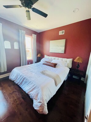 Een slaapkamer met een bed, een plafondventilator, een raam met gordijnen en een wandlamp.