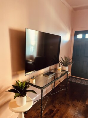 Een flatscreen televisie aan een muur bevestigd, een glazen salontafel met een plant, en een deur op de achtergrond.