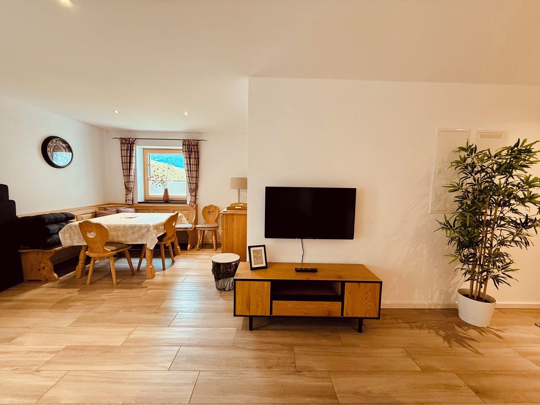 A living room with a TV, wooden furniture, a dining table, and a potted plant.