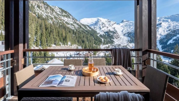 A wooden table with a magazine, a tray with pastries, and a glass of drink on a balcony with a mountain view. - Alps Flat With Heated Pool (Araches-la-Frasse)