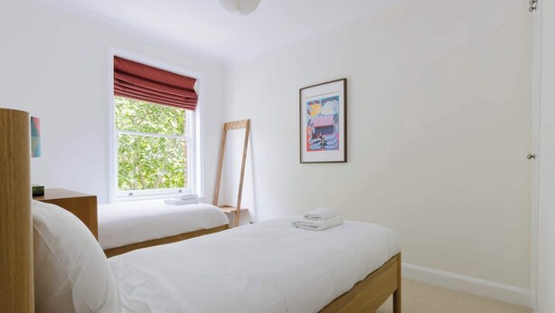 A bedroom with two beds, a window with red curtains, a framed picture on the wall, and a wooden bedside table. - Tasteful 2BDR Midcentury Modern Home (London)