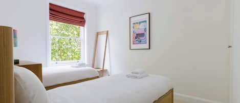 Un dormitorio con dos camas, una ventana con cortinas rojas, un cuadro en la pared y una mesita de noche de madera.