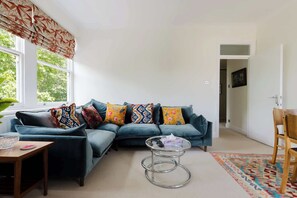 A living room with a sectional sofa, a round coffee table, a patterned rug, and a window with a view of greenery. - Tasteful 2BDR Midcentury Modern Home (London)