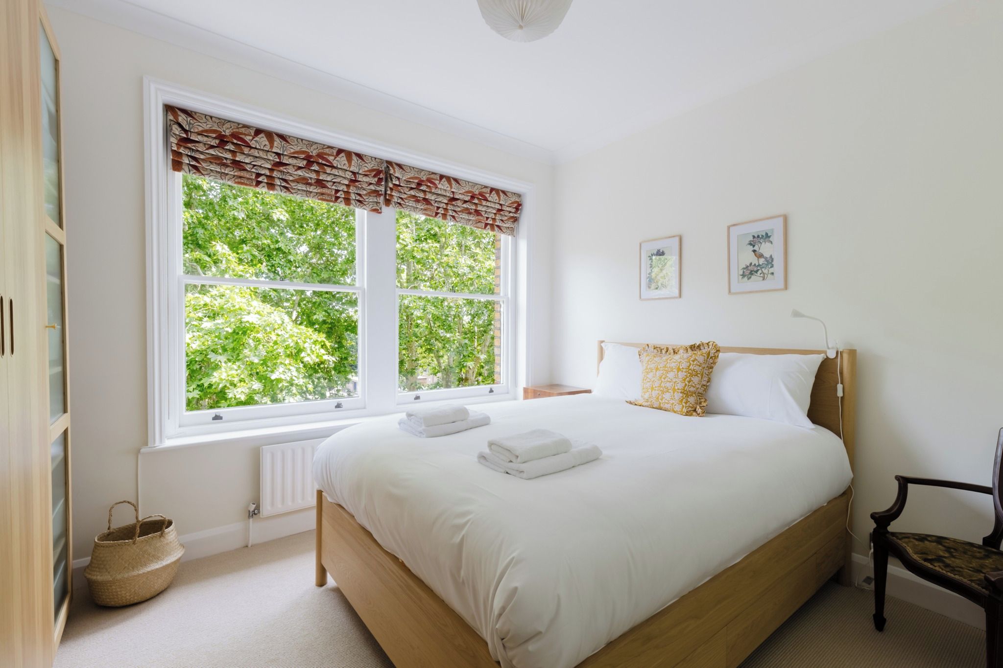 A bedroom with a large window, a bed with white linens, a wooden headboard, and two framed pictures on the wall.