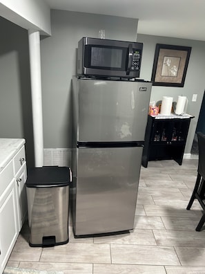 A kitchen with a stainless steel refrigerator, a microwave, a trash bin, and a small table with a picture frame. - Cozy 1-bedroom basement apartment suite in lovely Elmont with private entrance. (Elmont)