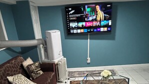 Una sala de estar con un televisor montado en la pared, un sofá con cojines estampados, una mesa de centro de vidrio con un jarrón de flores y un radiador.