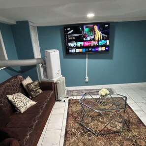 A living room with a TV mounted on the wall, a sofa with patterned cushions, a glass coffee table with a vase of flowers, and a radiator. - Cozy 1-bedroom basement apartment suite in lovely Elmont with private entrance. (Elmont)