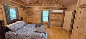 Un dormitorio de cabaña de madera con cama, dos ventanas, un equipo de aire acondicionado y una puerta.