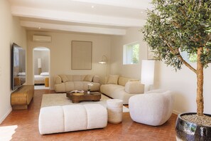 Ein modernes Wohnzimmer mit einer Eckcouch, Poufs, einem Couchtisch, einem großen Kübelbaum und einem Flachbildfernseher.
