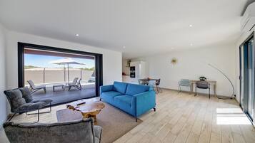 A spacious living area with a blue sofa, grey armchair, and a wooden coffee table. A large window offers a view of the outdoors.