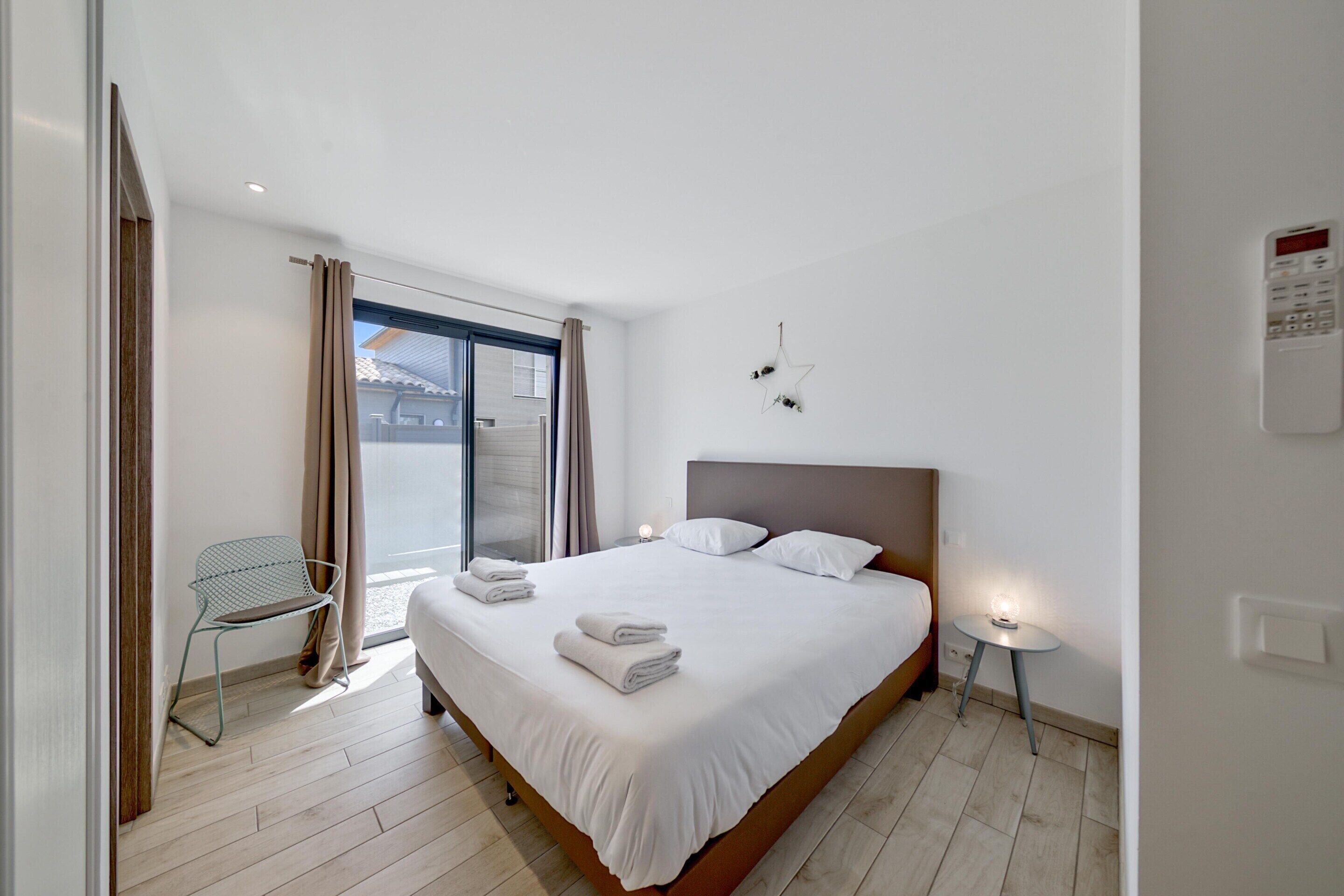 A modern hotel room with a large bed, a chair, a small table, and a window with curtains.