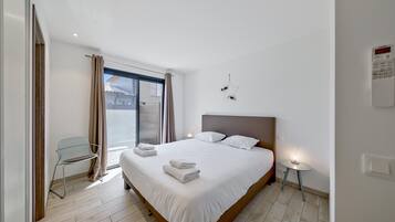 A modern hotel room with a large bed, a chair, a small table, and a window with curtains.