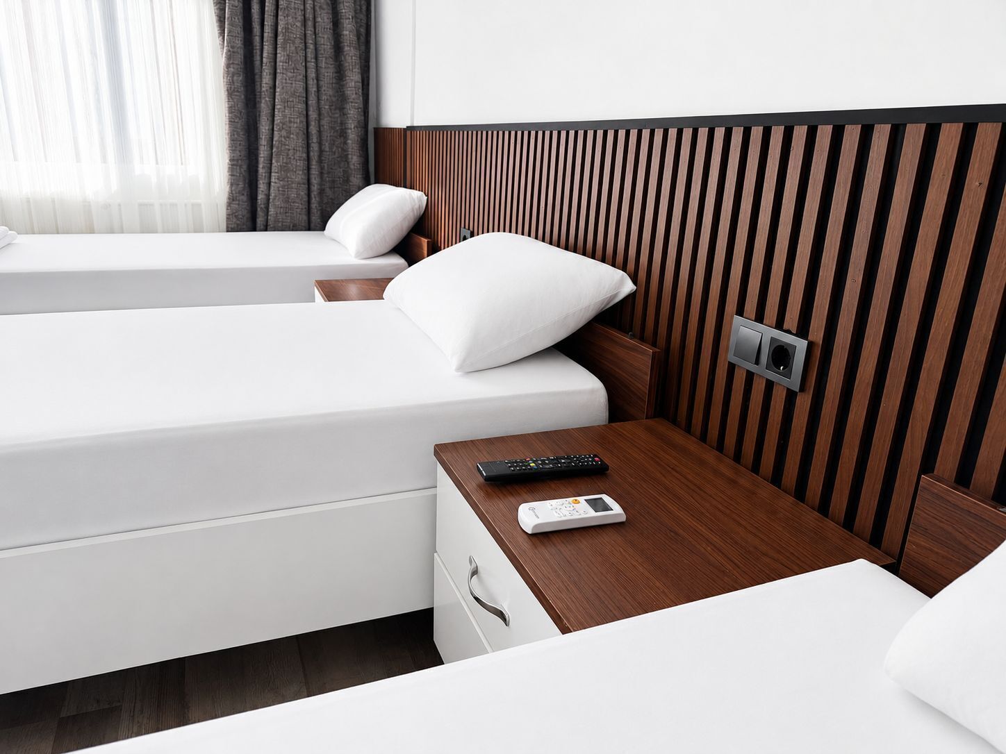 Quarto de hotel com duas camas, uma mesa de cabeceira de madeira, um controle remoto, um termostato e um interruptor de luz fixado na parede.