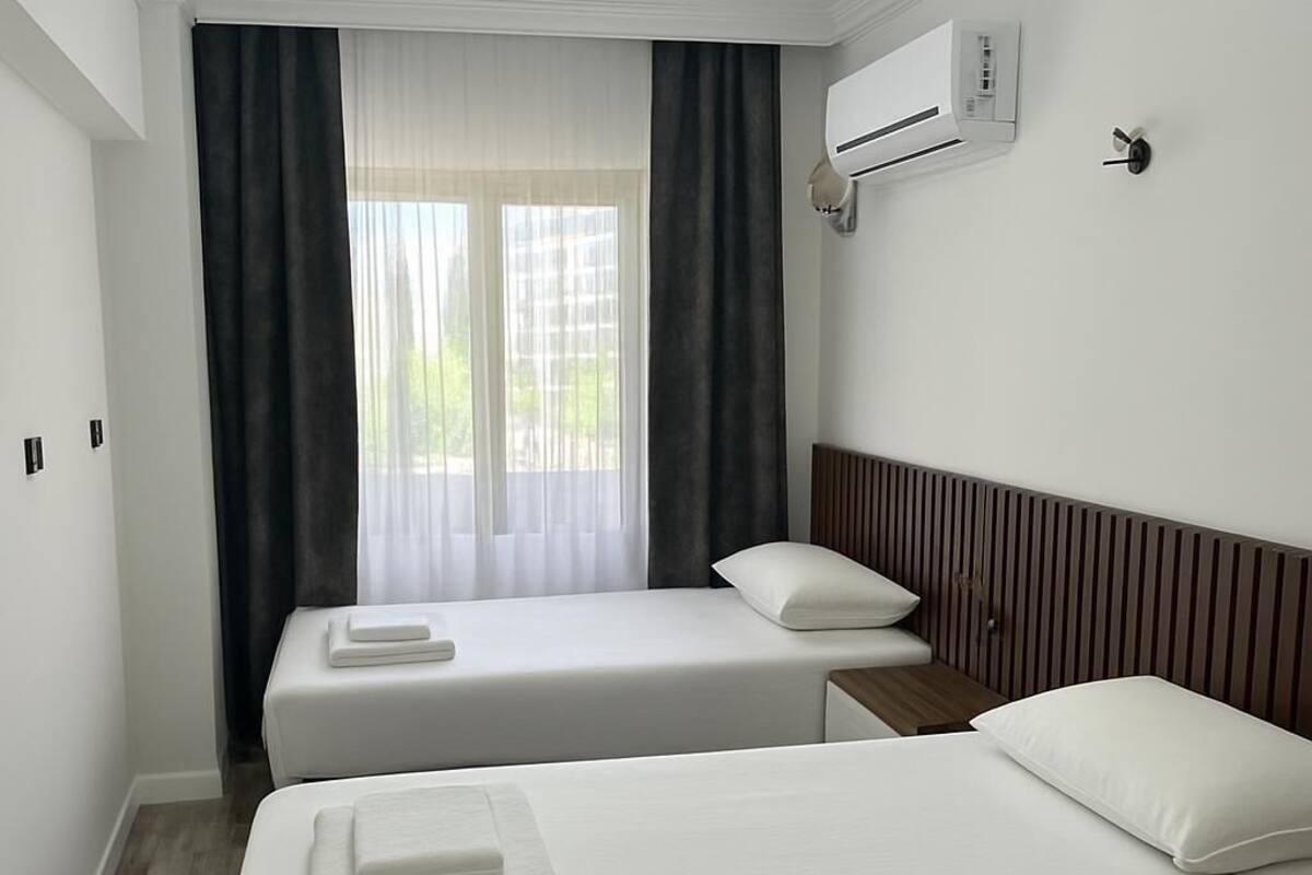 A hotel room with two beds, a window with curtains, an air conditioning unit, and a wall-mounted light.