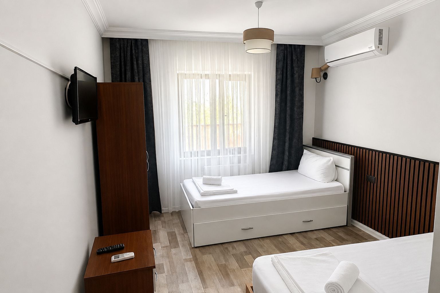 A hotel room with a bed, a TV mounted on the wall, a bedside table with a remote control, and an air conditioning unit on the wall.