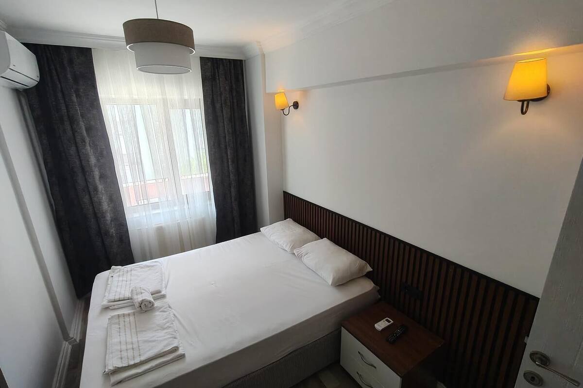 A hotel room with a bed, a nightstand, a lamp, an air conditioner, and a window with curtains.