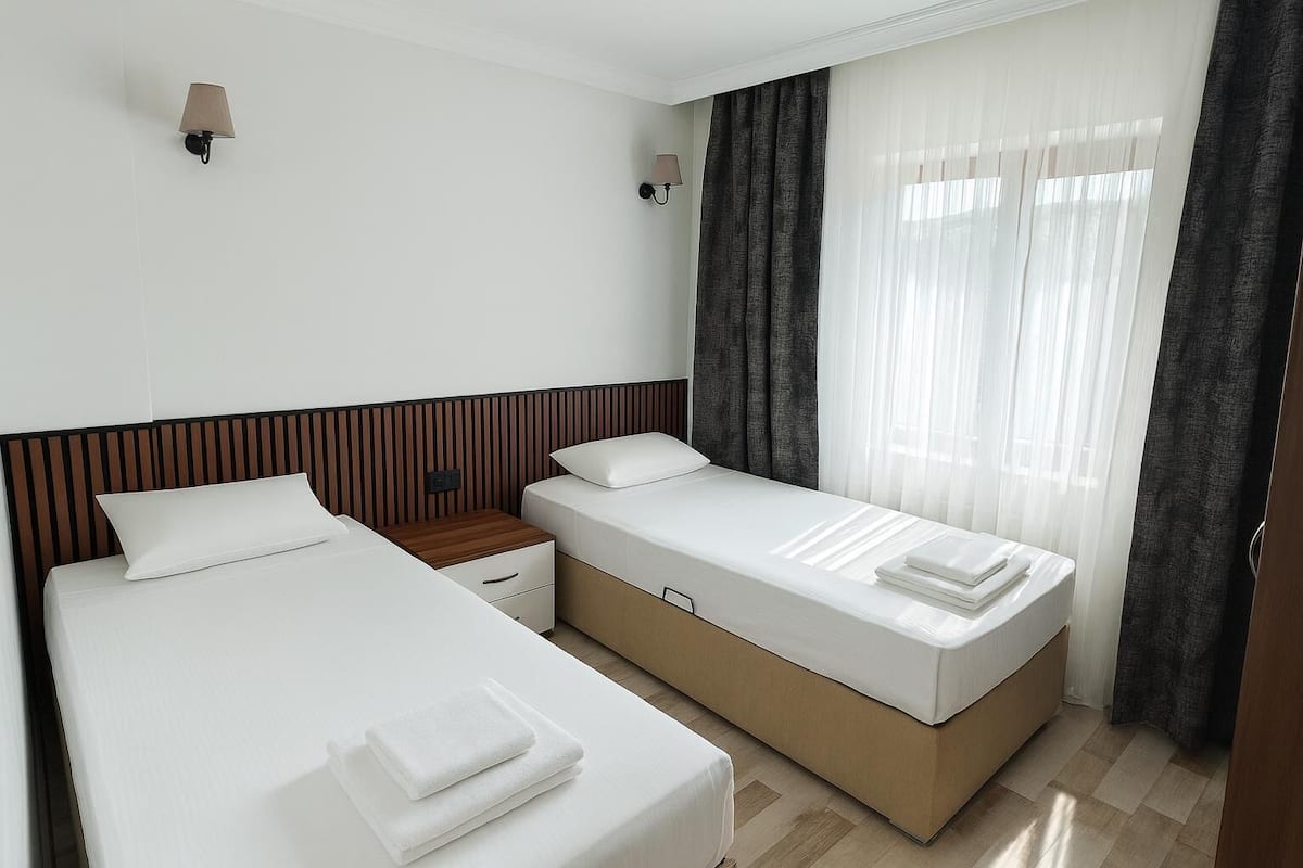 A hotel room with two beds, a nightstand, a wall-mounted lamp, and a window with curtains.