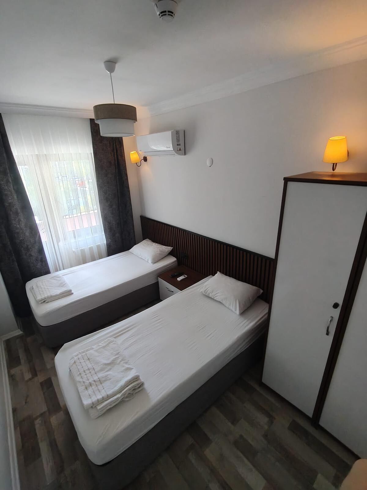 A hotel room with two single beds, a wardrobe, a bedside table with a lamp, and a window with curtains.