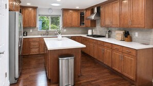 A modern kitchen with wooden cabinets, a central island, a stainless steel trash can, and a window with a view of greenery. - Bellevue 2323, Whole house, Fenced Yard (Bellevue)