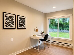 Ein Zimmer mit weißem Schreibtisch, schwarzem Stuhl, zwei gerahmten Blumenbildern und einem großen Fenster mit Blick auf Grünflächen.